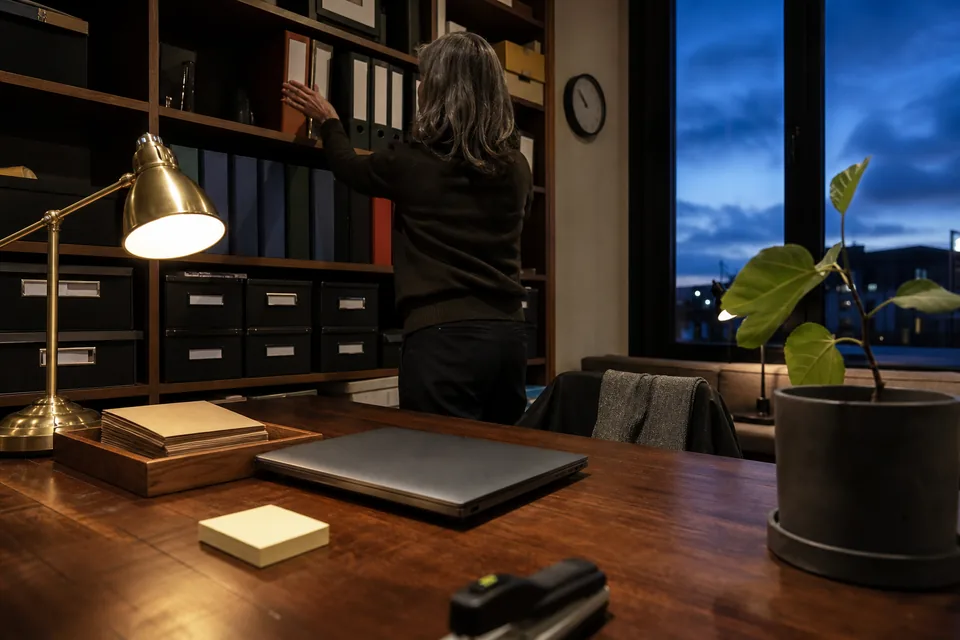 Agency finance desk at dusk with a lit brass lamp, ring binders on a shelf and a closed laptop on a walnut desk, a woman filing a binder