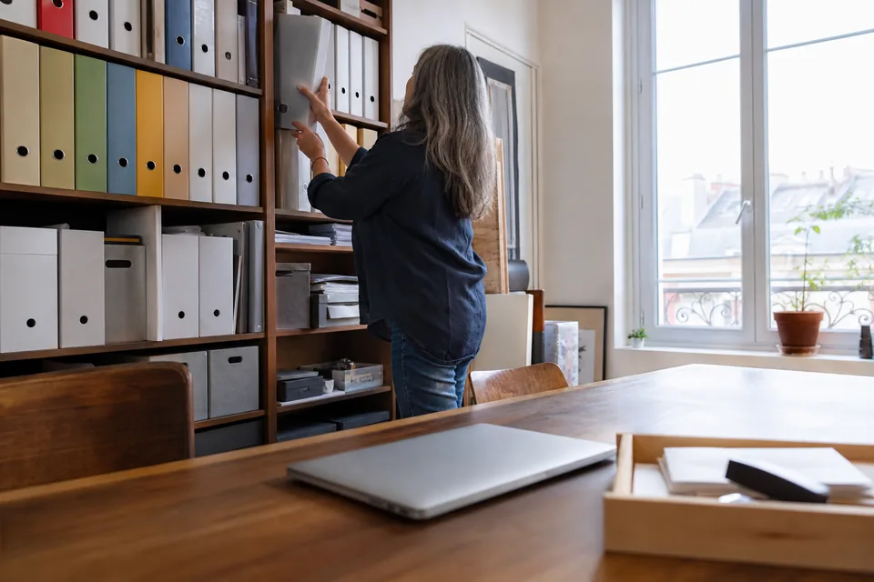 Agency back office in daylight, a woman filing a ring binder on a shelf of binders and archive boxes, a walnut desk with a closed laptop and a tray of envelopes in front