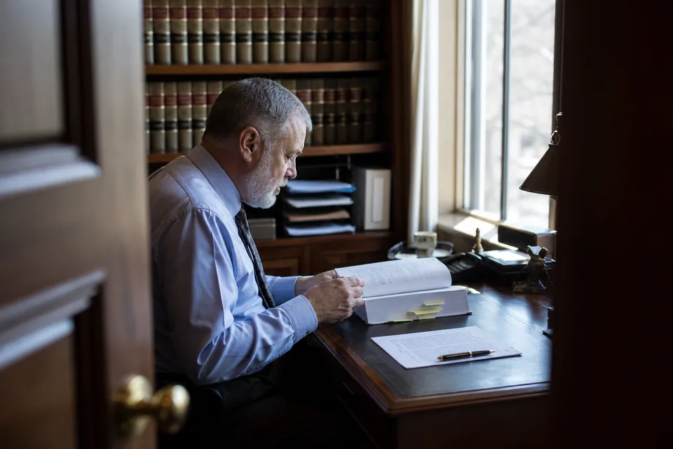 Compliance officer reading a bound policy binder at a window desk with sticky notes