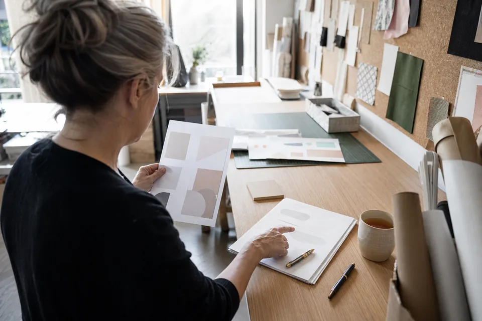 Studio owner at a plywood work counter comparing a printed report with a client contract, a pen and a signed proposal beside her, seen from behind her shoulder