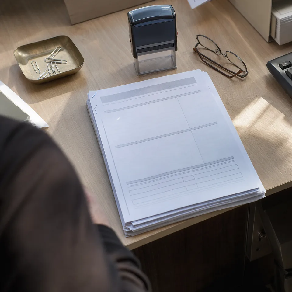 Close detail of a desk with a stack of stapled company filings, a stamp and reading glasses in daylight